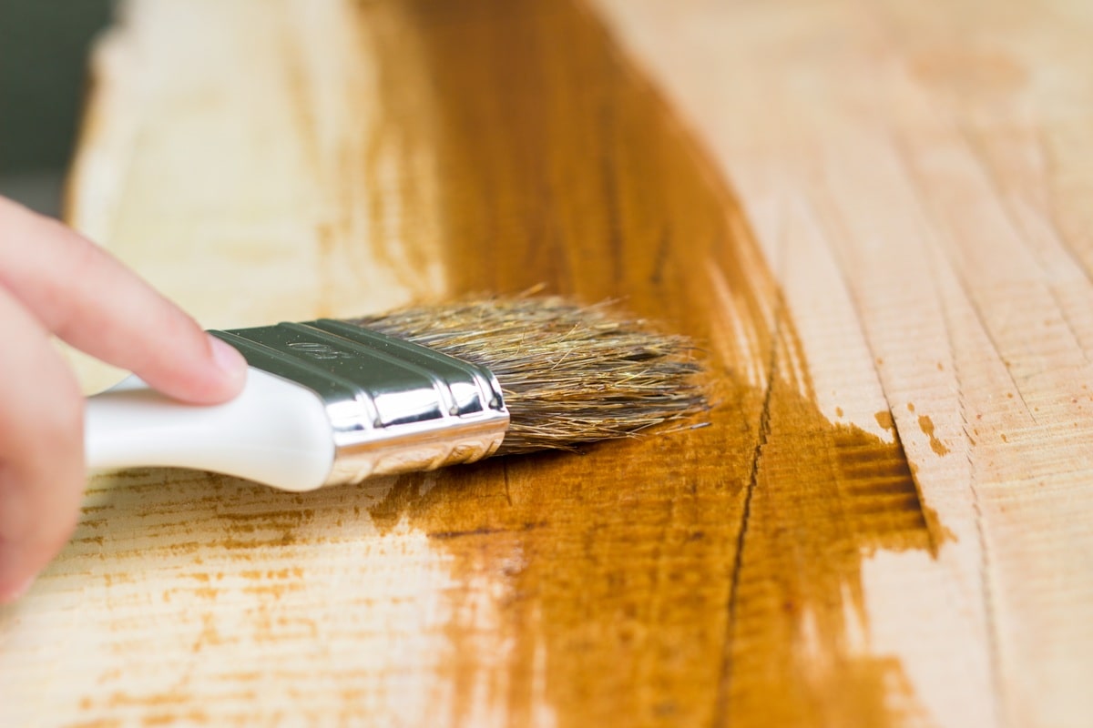 A imagem mostra uma mão aplicando stain com um pincel sobre uma superfície de madeira, destacando a textura e o brilho do acabamento.