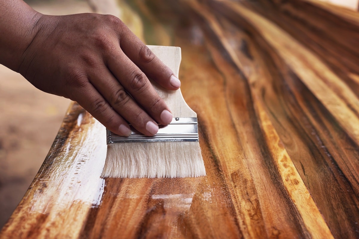 Homem aplicando acabamento em verniz na madeira com pincel, destacando a textura e brilho da superfície de um móvel. Importante observar as tendências dos fabricantes.